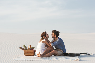 young couple hugging while sitting on blanket with basket of fruits and acoustic guitar on beach