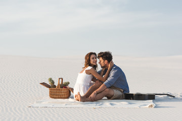 young couple embracing while sitting on blanket with basket of fruits and acoustic guitar on beach