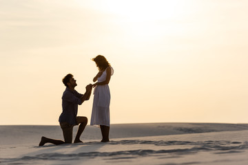 side view of young man doing marriage proposal to girlfriend on sandy beach at sunset