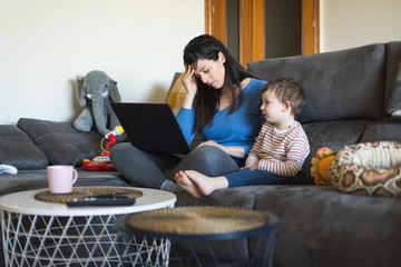 Tired woman working on laptop at home with her child. Single mother telecommuting.
