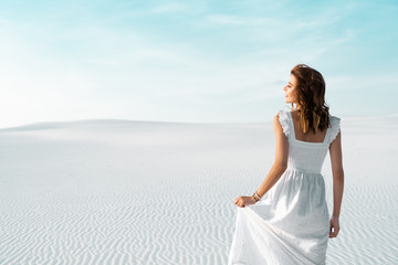 Fototapeta na wymiar back view of beautiful girl in white dress on sandy beach with blue sky