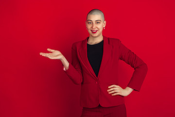 Showing, presenting. Portrait of young caucasian bald woman isolated on red studio background. Beautiful female model in jacket. Human emotions, facial expression, sales, ad concept. Freaky culture.