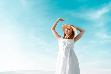 Fototapeta na wymiar low angle view of smiling beautiful girl in white dress and straw hat against blue sky