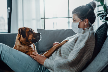 Woman wearing a protective face mask cuddles, plays with her dog at home because of the corona virus pandemic covid-19