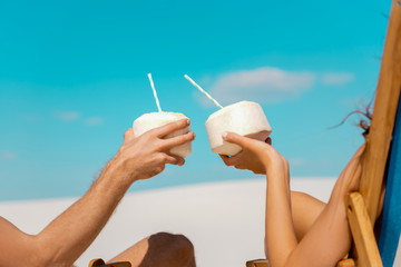 cropped view of young couple sitting in deck chairs with coconut drinks on sandy beach