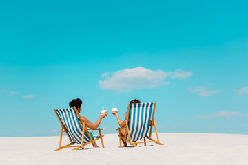 back view of young couple sitting in deck chairs with coconut drinks on sandy beach