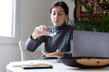 Young woman cleaning her hands with alcohol and tele working on her computer at home during quarantine because coronavirus.
