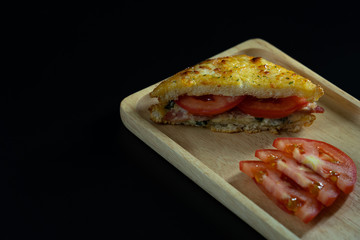 Baked croque monsieur spinach and ham sandwich with tomato on wood plate, isolated black background.