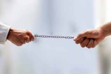 Shot of two hands on the end of a rusted metal chain pulling from both sides with blurred background and selective focus. Male hand playing tug of the war with a metal chain.