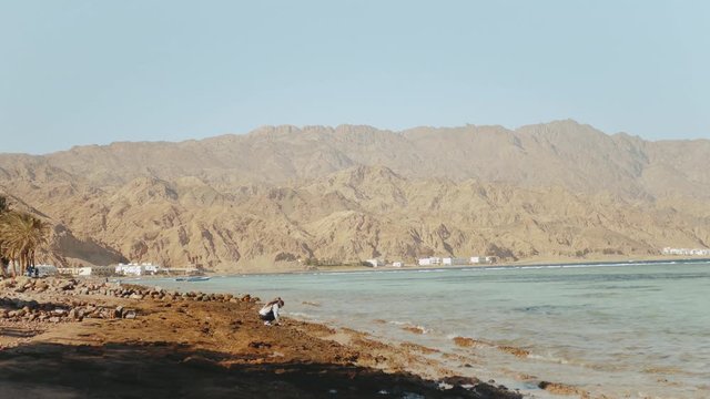 Pretty woman walking alone on stony shore beach near sea, the waves are breaking on the shore, Egypt Sinai mountain on the background, slow motion, 4k