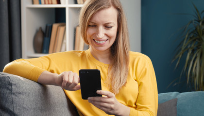 Lovely cheery blond mature woman scrolling through smartphone sitting and leaning arm on couch back