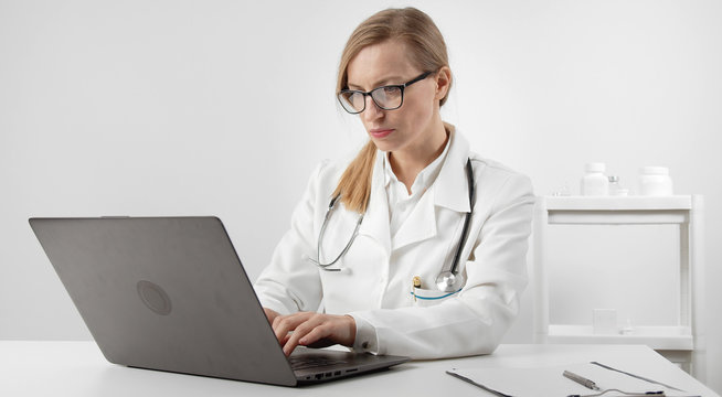Portrait of serious female doctor using laptop typing keyboard sitting at desk in clinic