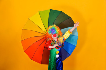 Clown child in a mask with a large colored umbrella on a yellow background creates a festive mood.