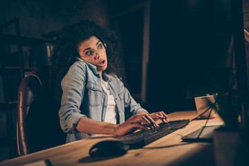 Fototapeta na wymiar Photo of pretty dark skin business lady look monitor work overtime talk telephone partner busy rushing typing text deadline wear specs casual shirt sit table chair night office indoors