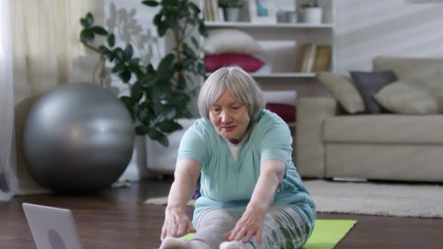 Tilt Down Shot Of Senior Woman Sitting On Mat On The Floor In Living Room And Trying To Touch Her Toes While Following Stretching Lesson On Laptop