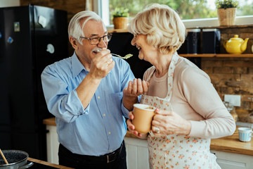 Happy senior man having fun while feeding his wife in the kitchen.