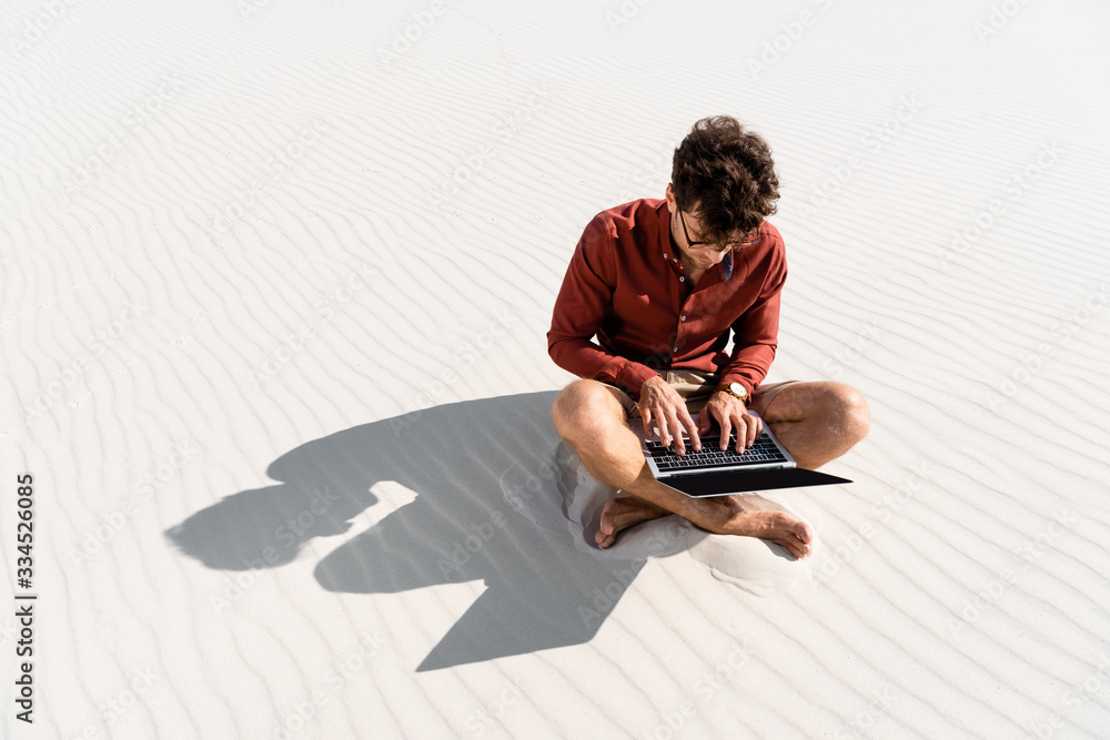 Wall mural young freelancer sitting on sandy beach with laptop