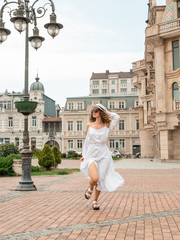 Girl female in a white long dress, with a backpack, holding hat walking near the clock. Back view of attractive and happy girl in old cozy streets of Europe. Pretty sexy fashion model in cityscape. Li