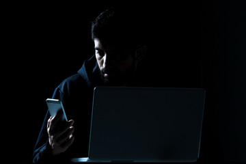 Dramatic shot of hacker using laptop and smartphone in dark room