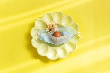 Fototapeta na wymiar One egg in plate and medical mask on yellow background. Concept of canceling the Easter holiday during the global pandemic. Flat lay, top view.