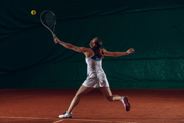 Young caucasian professional sportswoman playing tennis on sport court background. Training, practicing in motion, action. Power and energy. Movement, ad, sport, healthy lifestyle concept. Front view.