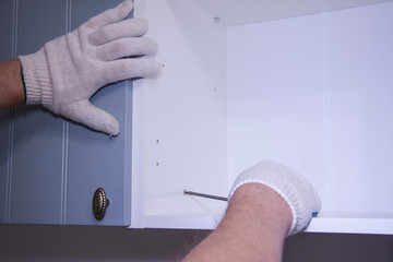 Repair and installation of furniture in the room. Male working hands in white gloves with a screwdriver. Assembly of kitchen cabinets. Details and units of furniture with bolts and fixtures.