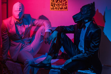 two guys, one in a black suit and a dog mask, one in a white suit with a white latex mask, is sitting on a stretcher