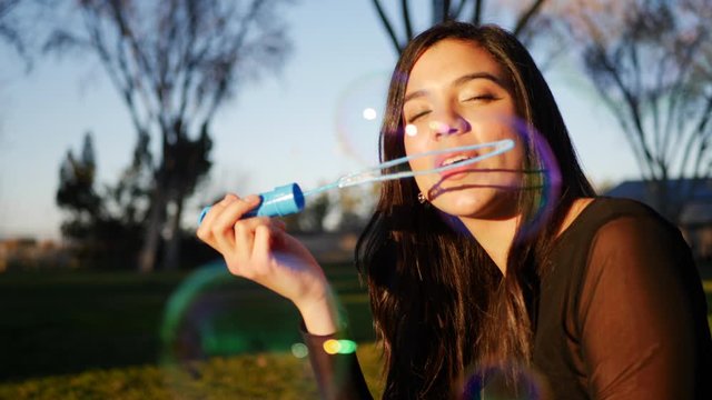 Beautiful young hispanic woman blowing dreamy and colorful bubbles while smiling and looking happy outdoors at sunset.