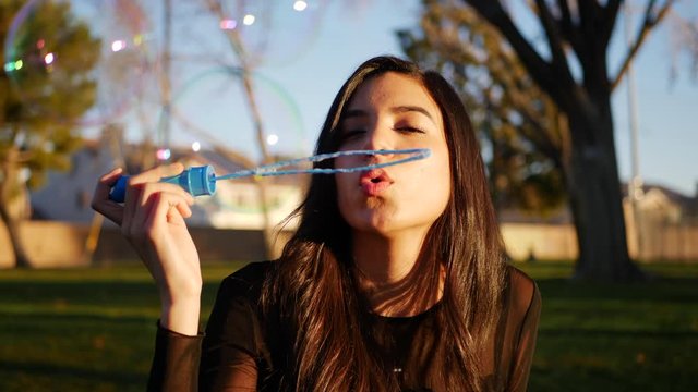 Attractive hispanic woman blowing dreamy and colorful bubbles while looking nostalgic and serious outdoors at sunset.