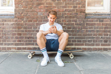 Naklejka premium Man sitting on a skateboard and using a phone in London - portrait against a wall of a young man relaxing and staying connected to social media - Lifestyle and technology concepts