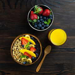 Healthy breakfast. A wooden spoon and a bowl of granola, fruits and glass of orange juice. Blueberry and banana smoothie dressing, decorated with strawberries, mangoes. Flatlay on dark background.