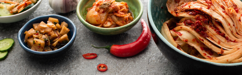 panoramic shot of kimchi in bowls, chili pepper and garlic on concrete surface