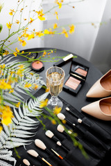 Beautiful morning of a bride. Yellow flowers composition and fern, makeup brushes, highlighter, biege shoes and a glass of champagne on a black wooden table. For wedding publications and advertising.