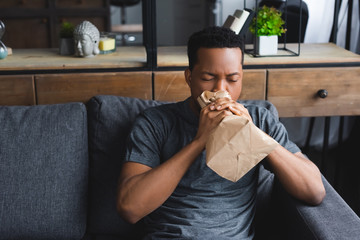 stressed african american man breathing with paper bag while having panic attack at home