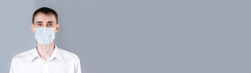 Banner. Man in a medical mask. A handsome dark-haired man in a white medical uniform, shirt and medical mask  looking at camera on a gray background, on the right there is an empty space.  Coronavirus