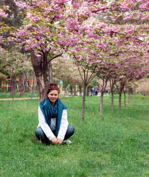 Young happy smiling woman with jeans pants, white sweater and blue scarf sits on the grass under a blossoming pink sakura tree in a park in the city of Kiev, Ukraine