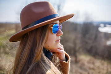A girl in a brown coat, hat and glasses walks in the park with a lake under the bright sun.