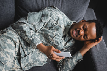 lonely african american soldier crying while holding army badge and smartphone, suffering from PTSD at home