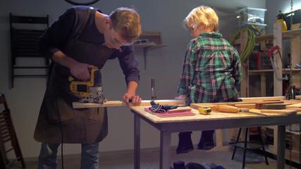 Boy sits on board, man screws a nail into a board on a table