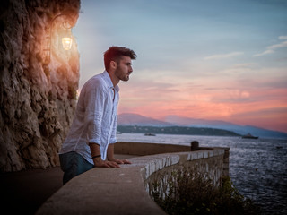 Side view of male in white shirt leaning on border and admiring sea while standing on embankment near rough cliff with lantern against sundown sky