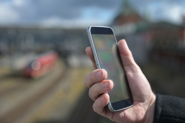 Hand of a man with a mobile phone, station, railroad tracks and train blurred in the background, concept for tracking and ticket apps during traffic shorting due to coronavirus pandemic, copy space