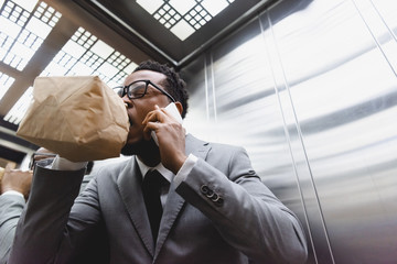 stressed african american businessman talking on smartphone while breathing with paper bag and suffering from panic attack in elevator - obrazy, fototapety, plakaty