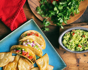 Overhead view of several tacos on a square blue plate. Shown on wooden table with fresh toppings including cilantro and guacamole. 
