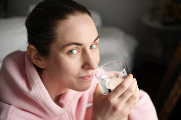 Beautiful woman wearing home clothing looking aside, while sitting in chair in living room and holding glass of water.