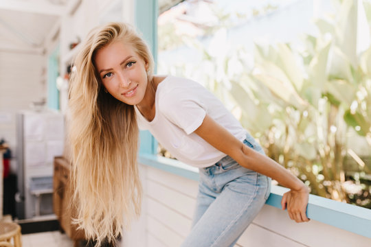 Graceful lady with long blonde hair having fun in weekend and posing with pleasure. Enthusiastic caucasian woman with tanned skin laughing while standing near window.