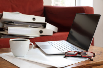 Laptop, coffee cup and glasses on a living room table and a lot of ring binders on the sofa behind,...