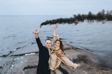 Stylish newlyweds hug and hold a pyrotechnic device with black smoke against the background of the river, sea and sky, standing on the pier. Photography, concept.