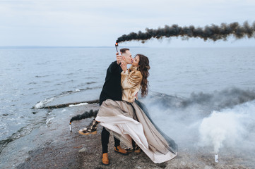 Stylish newlyweds hug, kiss and hold a pyrotechnic device with black smoke against the background...