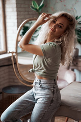Attractive young woman with blonde curly hair wearing casual clothes posing in a bright apartment leaning on wooden table and holding her hand up