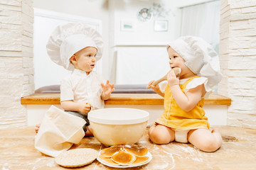 Restless cute caucasian grimy two-year-old children brother and sister are sitting on the kitchen table looking at each other with cutlery with fritters. Concept of passionate children at home